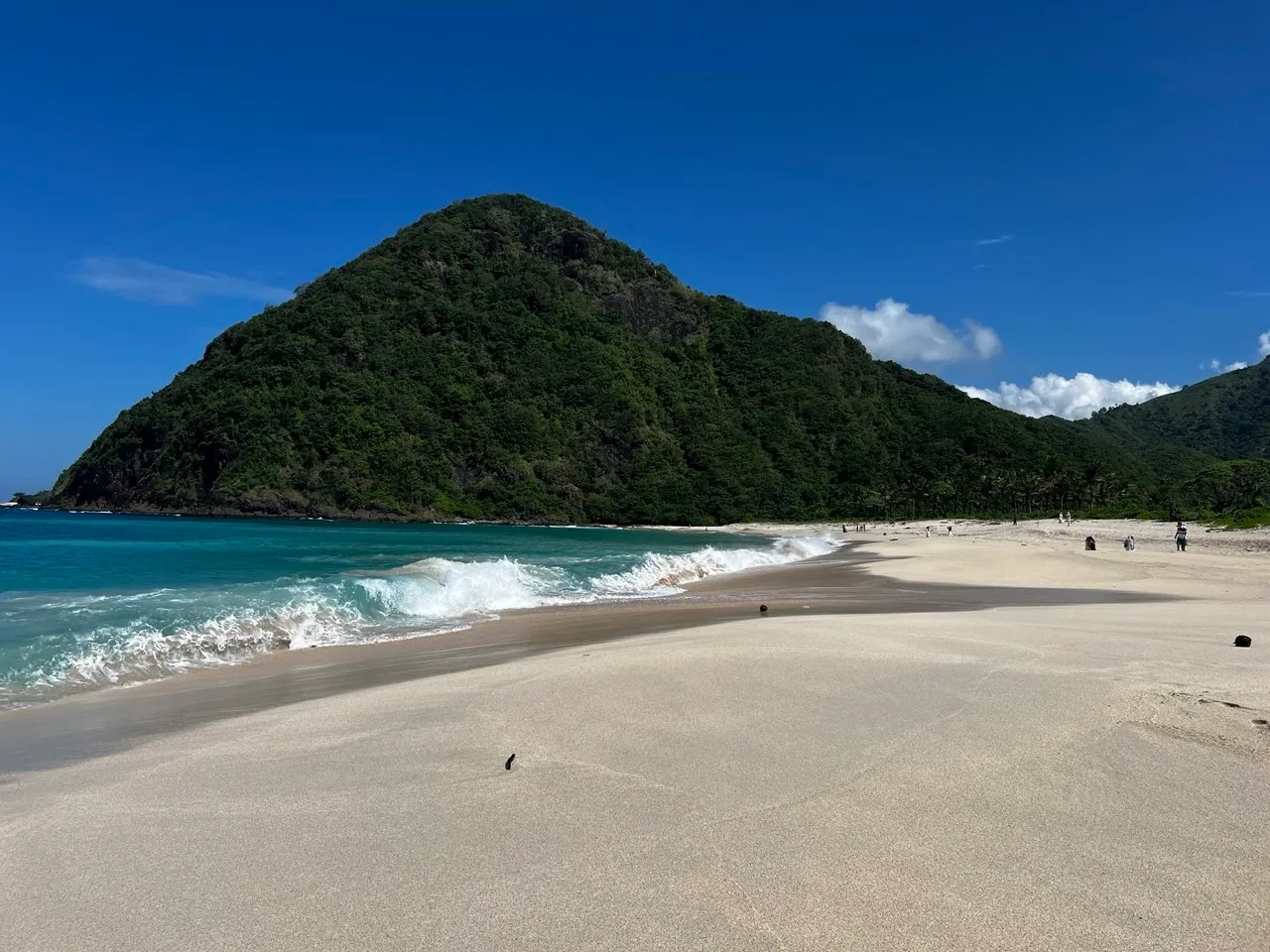 Selong Belanak beach: a white-sand bay below a green conical headland with gentle surf, South Lombok.